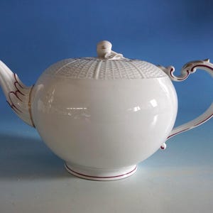 Può includere: Una teiera in porcellana bianca con corpo rotondo, coperchio strutturato e beccuccio curvo. Il manico e il beccuccio sono impreziositi da dettagli bordeaux e oro. La teiera poggia su una base bordata di bordeaux, un classico per il servizio da tè.