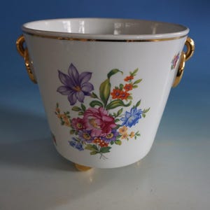 Op de afbeelding: Witte keramische plantenpot met gouden rand en bloemenmotief. De pot heeft twee gouden handvatten en drie gouden voetjes.