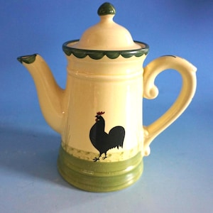 Può includere: Teiera in ceramica color crema con un'illustrazione di gallo nero. La teiera ha un bordo smerlato verde, un coperchio abbinato e un manico curvo. Anche la base e il beccuccio sono accentuati in verde. Un'aggiunta affascinante a qualsiasi cucina.