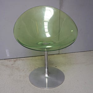 Puede incluir: Una moderna silla de cristal verde con un asiento redondeado en forma de cuenco. El asiento está sostenido por un tallo de metal cepillado y una base circular. La silla tiene un diseño minimalista y una estética retro.