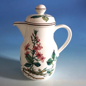 Pode incluir: Jarra de café de cerâmica branca com tampa e alça. A jarra é decorada com um desenho floral com flores cor-de-rosa, folhas verdes e detalhes castanhos. A tampa tem um pequeno botão e uma borda castanha. A jarra tem um bico.