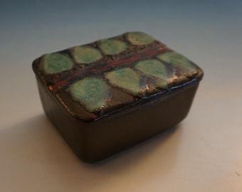 May include: A small, rectangular ceramic box with a lid. The lid is decorated with a pattern of green and brown leaves. The box is a dark brown color.