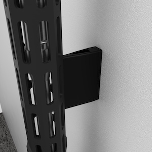 May include: Black metal shelving unit with a slotted design, mounted on a white wall. A black bracket is attached to the wall, supporting the shelving. The image shows a close-up of the corner of the shelving unit.