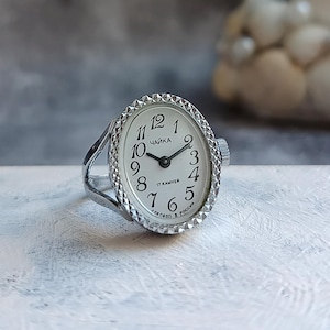 May include: A silver-toned ring watch featuring an oval face with a white dial, black Arabic numerals, and thin black hands. The bezel has a textured, diamond-cut pattern. The dial displays the text "ЧАЙКА", "17 КАМНЕЙ", and "СДЕЛАНО В РОССИИ".