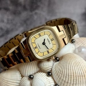 May include: A gold-toned vintage wristwatch with a rectangular face and a matching link band. The watch face is cream-colored with black hands and Roman numerals. The watch is displayed on a decorative arrangement of seashells and pearls.