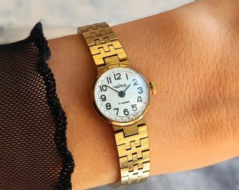 May include: A gold-tone analog wristwatch with a round white face, black Arabic numerals, and a textured metal link bracelet. The watch dial features the Cyrillic text "ЧАЙКА" and "17 КАМНЕЙ" in black print, with a small gold crown on the side.