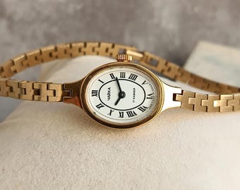 May include: A gold-tone wristwatch with an oval-shaped case and a matching link bracelet. The white dial features black Roman numeral hour markers and black hands. The text "ЧАЙКА" and "17 КАМНЕЙ" is printed on the dial face.