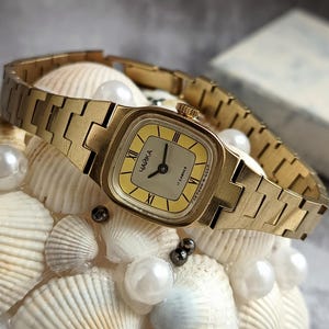 Pu&ograve; includere: Un orologio da polso vintage color oro con quadrante quadrato e quadrante giallo chiaro. L'orologio presenta numeri romani e la scritta "ЧАЙКА" sul quadrante. &Egrave; esposto su un letto di conchiglie e perle.