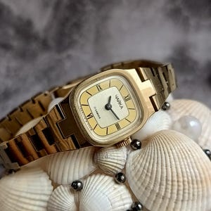 Pu&ograve; includere: Un orologio da polso vintage color oro con quadrante rettangolare e cinturino abbinato. Il quadrante dell'orologio &egrave; color crema con lancette nere e numeri romani. L'orologio &egrave; etichettato "ЧАЙКА". L'orologio &egrave; esposto su un letto di conchiglie e perle.
