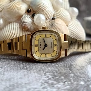 Pu&ograve; includere: Un orologio da polso vintage color oro con quadrante rettangolare e cinturino abbinato. Il quadrante &egrave; giallo chiaro con numeri romani neri e la parola cirillica "ЧАЙКА". L'orologio &egrave; ambientato su uno sfondo di conchiglie e perle.