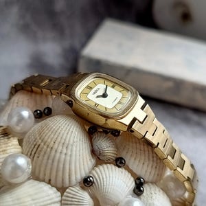 Pu&ograve; includere: Un orologio da polso dorato con quadrante rettangolare e cinturino abbinato. Il quadrante dell'orologio &egrave; color crema con lancette nere e numeri romani. L'orologio &egrave; esposto su un letto di conchiglie e perle.