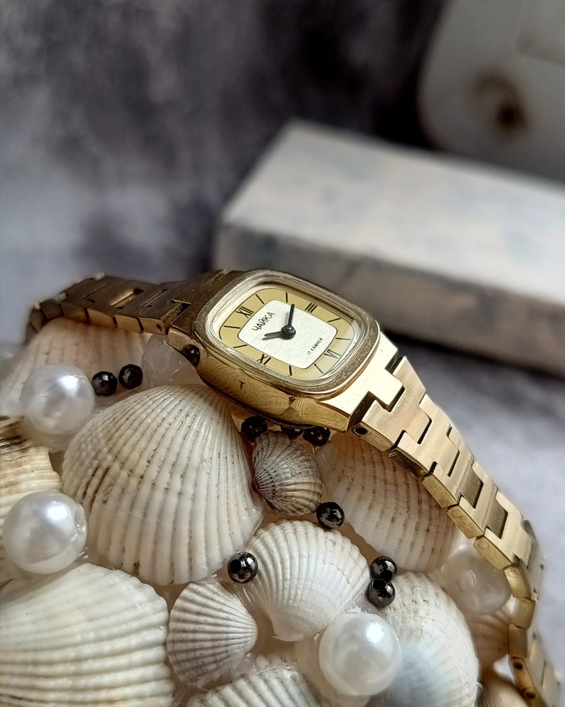 Pu&ograve; includere: Un orologio da polso dorato con quadrante rettangolare e cinturino abbinato. Il quadrante dell'orologio &egrave; color crema con lancette nere e numeri romani. L'orologio &egrave; esposto su un letto di conchiglie e perle.