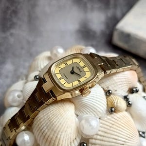 Pu&ograve; includere: Un orologio da polso vintage color oro con quadrante rettangolare e cinturino in metallo abbinato. Il quadrante dell'orologio &egrave; color oro chiaro con numeri romani neri e il marchio "ЧАЙКА". L'orologio &egrave; esposto su un letto di conchiglie e perle.