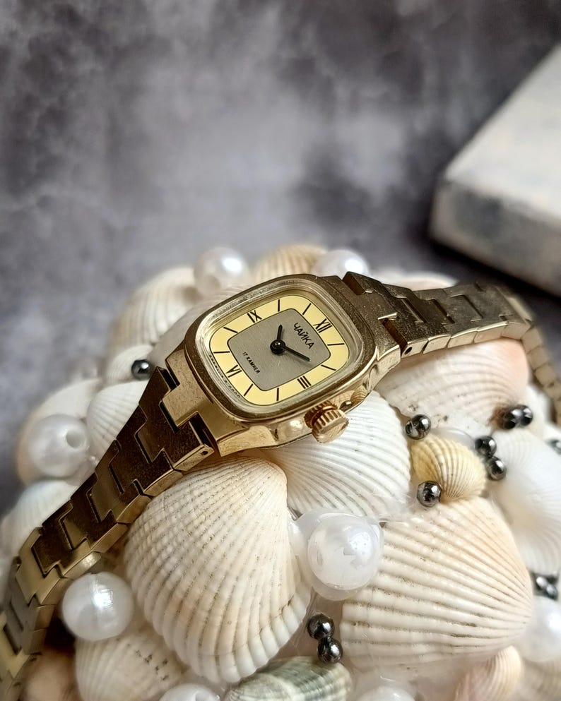 Pu&ograve; includere: Un orologio da polso vintage color oro con quadrante rettangolare e cinturino in metallo abbinato. Il quadrante dell'orologio &egrave; color oro chiaro con numeri romani neri e il marchio "ЧАЙКА". L'orologio &egrave; esposto su un letto di conchiglie e perle.