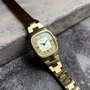 Pu&ograve; includere: Un orologio da polso vintage color oro con quadrante rettangolare. Il quadrante &egrave; color crema con lancette nere e numeri romani. Il cinturino dell'orologio &egrave; in tinta oro. Sul quadrante &egrave; stampata la parola "ЧАЙКА".