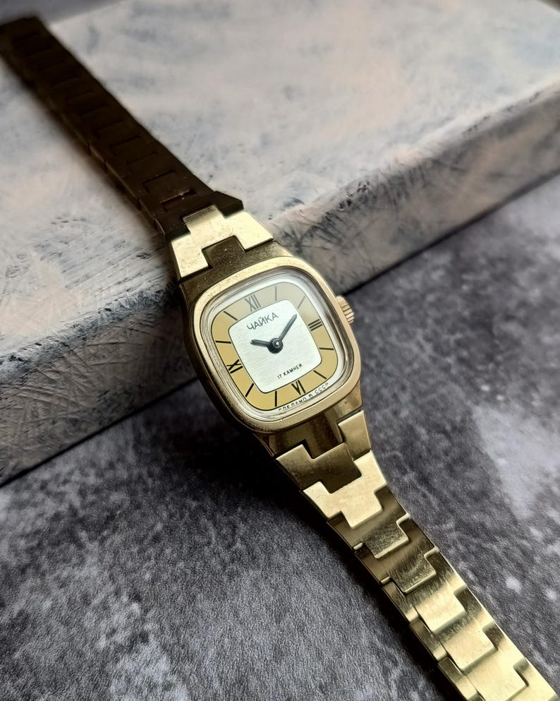 Pu&ograve; includere: Un orologio da polso vintage color oro con quadrante rettangolare. Il quadrante &egrave; color crema con lancette nere e numeri romani. Il cinturino dell'orologio &egrave; in tinta oro. Sul quadrante &egrave; stampata la parola "ЧАЙКА".