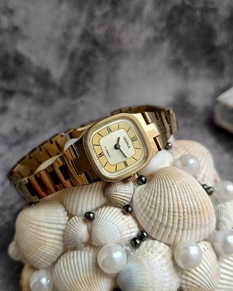 Pu&ograve; includere: Un orologio da polso vintage color oro con quadrante rettangolare e cinturino abbinato. Il quadrante dell'orologio &egrave; color crema con lancette nere e numeri romani. L'orologio &egrave; etichettato "ЧАЙКА". L'orologio &egrave; esposto su un letto di conchiglie e perle.