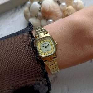Pu&ograve; includere: Un orologio da polso color oro con quadrante quadrato e quadrante giallo chiaro. L'orologio ha un cinturino a maglie e la parola "ЧАЙКА" &egrave; stampata sul quadrante. L'orologio &egrave; su un polso.