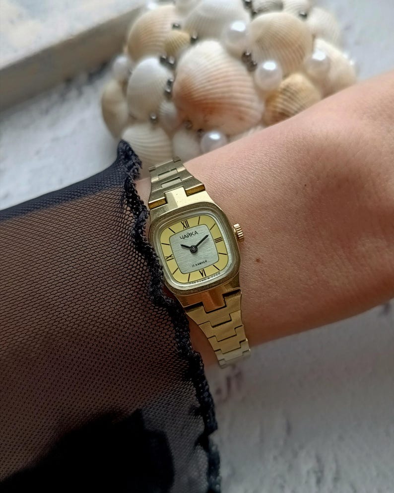 Pu&ograve; includere: Un orologio da polso color oro con quadrante quadrato e quadrante giallo chiaro. L'orologio ha un cinturino a maglie e la parola "ЧАЙКА" &egrave; stampata sul quadrante. L'orologio &egrave; su un polso.