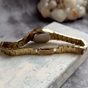 Pu&ograve; includere: Un orologio da polso color oro con quadrante rettangolare e cinturino a maglie. L'orologio &egrave; appoggiato su una superficie bianca e grigia. Il cinturino dell'orologio &egrave; lungo circa 15 cm.