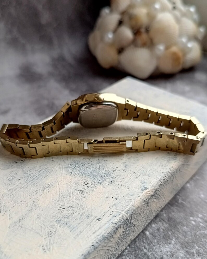 Pu&ograve; includere: Un orologio da polso color oro con quadrante rettangolare e cinturino a maglie. L'orologio &egrave; appoggiato su una superficie bianca e grigia. Il cinturino dell'orologio &egrave; lungo circa 15 cm.