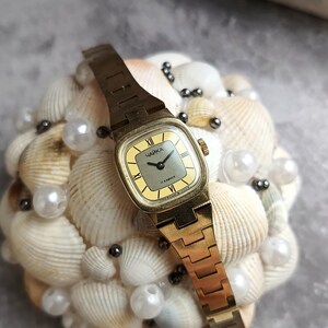Pu&ograve; includere: Un orologio da polso vintage color oro con quadrante quadrato e cinturino dorato. Il quadrante &egrave; giallo chiaro con numeri romani neri e la parola cirillica "Чайка". L'orologio &egrave; esposto su un letto di conchiglie e perle.