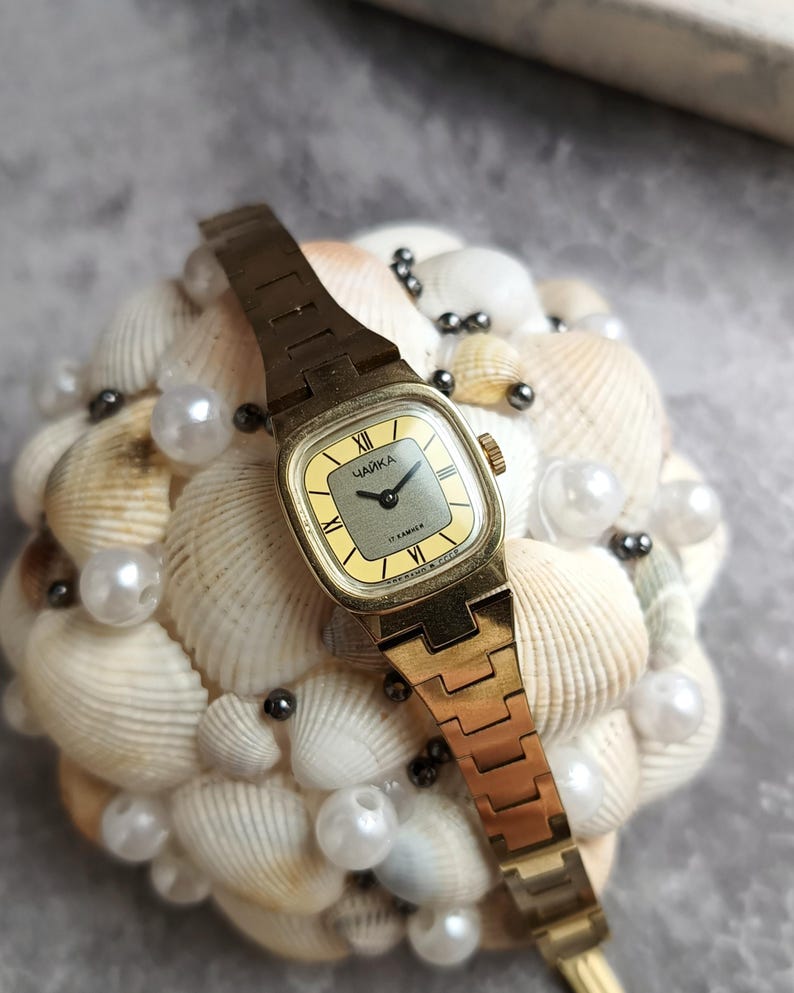 Pu&ograve; includere: Un orologio da polso vintage color oro con quadrante quadrato e cinturino dorato. Il quadrante &egrave; giallo chiaro con numeri romani neri e la parola cirillica "Чайка". L'orologio &egrave; esposto su un letto di conchiglie e perle.