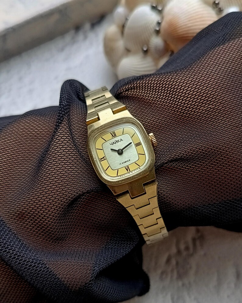 Pu&ograve; includere: Un orologio da polso vintage color oro con quadrante quadrato e cinturino abbinato. Il quadrante &egrave; giallo chiaro con lancette nere e numeri romani. La parola "ЧАЙКА" &egrave; stampata sul quadrante.