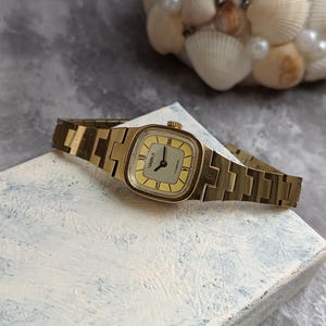 Pu&ograve; includere: Un orologio da polso vintage color oro con quadrante rettangolare e cinturino in metallo abbinato. Il quadrante &egrave; giallo chiaro con numeri romani neri e la scritta "ЧАЙКА" stampata. L'orologio &egrave; appoggiato su una superficie strutturata bianca e blu.