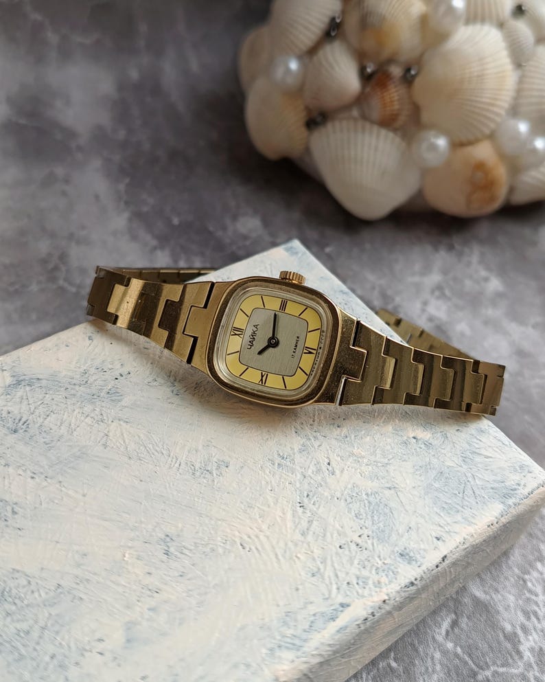 Pu&ograve; includere: Un orologio da polso vintage color oro con quadrante rettangolare e cinturino in metallo abbinato. Il quadrante &egrave; giallo chiaro con numeri romani neri e la scritta "ЧАЙКА" stampata. L'orologio &egrave; appoggiato su una superficie strutturata bianca e blu.