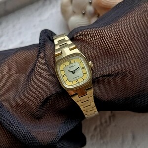 Pu&ograve; includere: Un orologio da polso vintage color oro con quadrante quadrato e cinturino abbinato. Il quadrante &egrave; color oro chiaro con lancette nere e numeri romani. L'orologio ha la scritta "ЧАЙКА" sul quadrante.