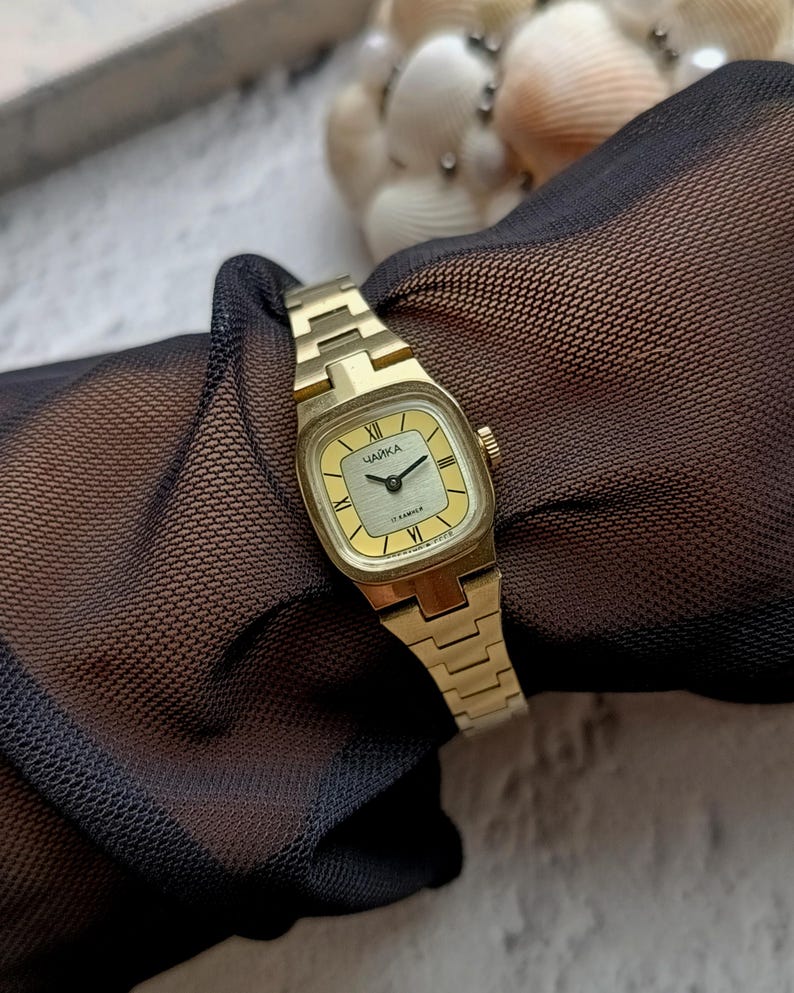 Pu&ograve; includere: Un orologio da polso vintage color oro con quadrante quadrato e cinturino abbinato. Il quadrante &egrave; color oro chiaro con lancette nere e numeri romani. L'orologio ha la scritta "ЧАЙКА" sul quadrante.