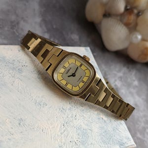 Pu&ograve; includere: Un orologio da polso vintage color oro con quadrante rettangolare e cinturino a maglie abbinato. Il quadrante dell'orologio &egrave; giallo chiaro con numeri romani neri e la scritta "ЧАЙКА" stampata sopra. L'orologio &egrave; appoggiato su una superficie bianca.