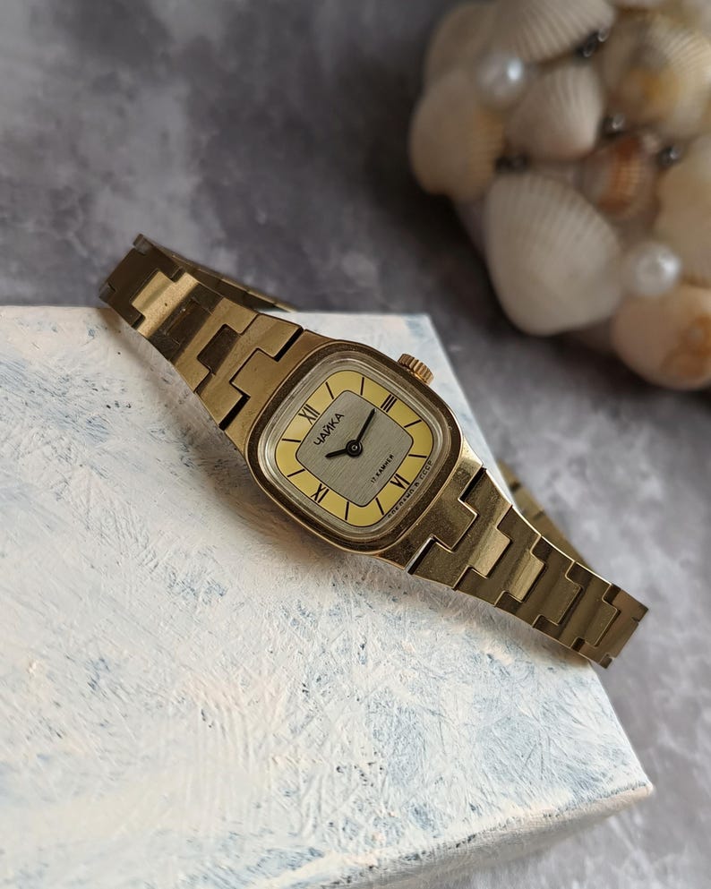 Pu&ograve; includere: Un orologio da polso vintage color oro con quadrante rettangolare e cinturino a maglie abbinato. Il quadrante dell'orologio &egrave; giallo chiaro con numeri romani neri e la scritta "ЧАЙКА" stampata sopra. L'orologio &egrave; appoggiato su una superficie bianca.