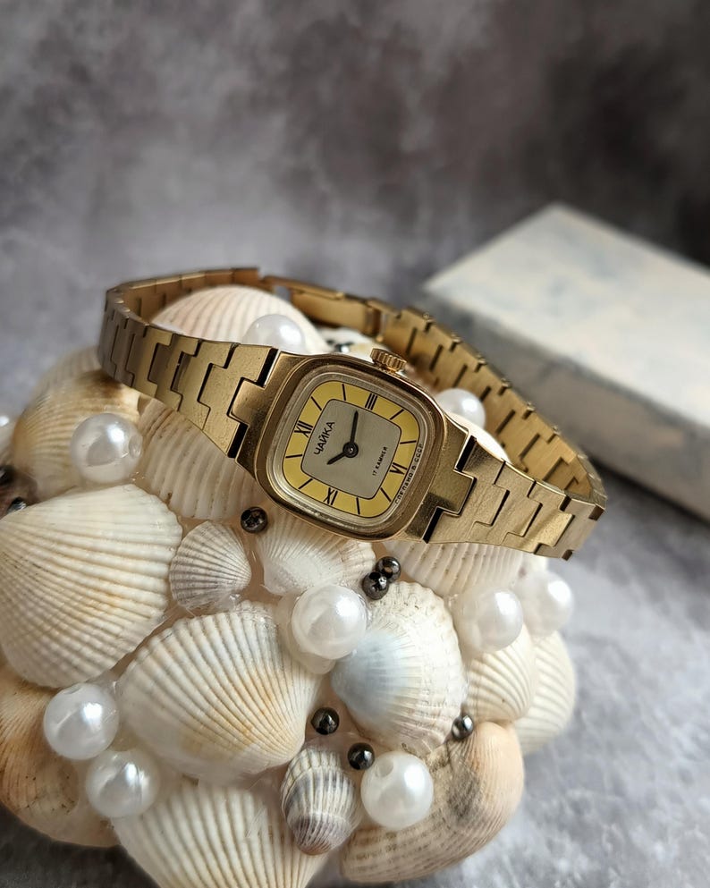 Pu&ograve; includere: Un orologio da polso vintage color oro con quadrante quadrato e quadrante giallo chiaro. L'orologio presenta numeri romani e la scritta "ЧАЙКА" sul quadrante. &Egrave; esposto su un letto di conchiglie e perle.