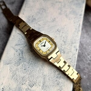 Pu&ograve; includere: Un orologio da polso vintage color oro con quadrante rettangolare color crema e cinturino a maglie abbinato. Il quadrante dell'orologio presenta numeri romani neri e il testo cirillico "ЧАЙКА". L'orologio ha un movimento a 17 rubini.