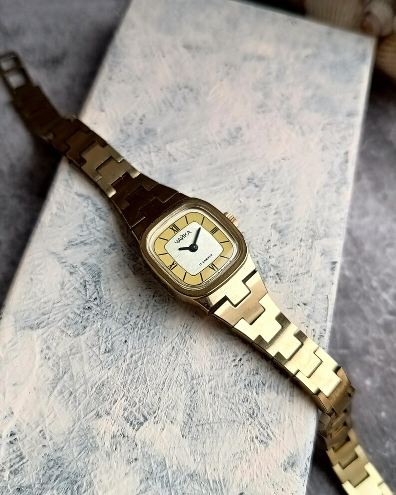Pu&ograve; includere: Un orologio da polso vintage color oro con quadrante rettangolare color crema e cinturino a maglie abbinato. Il quadrante dell'orologio presenta numeri romani neri e il testo cirillico "ЧАЙКА". L'orologio ha un movimento a 17 rubini.