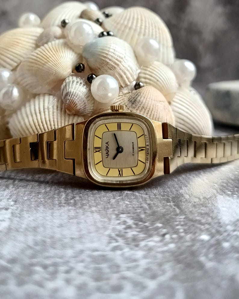 Pu&ograve; includere: Un orologio da polso vintage color oro con quadrante rettangolare e cinturino abbinato. Il quadrante &egrave; giallo chiaro con numeri romani neri e la parola cirillica "ЧАЙКА". L'orologio &egrave; ambientato su uno sfondo di conchiglie e perle.