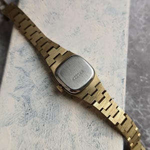Pu&ograve; includere: Un orologio da polso vintage color oro con un quadrante rettangolare color argento. Il cinturino dell'orologio &egrave; composto da maglie in metallo color oro. L'orologio ha il numero "413568" inciso sul retro. L'orologio &egrave; posto su uno sfondo chiaro e strutturato.