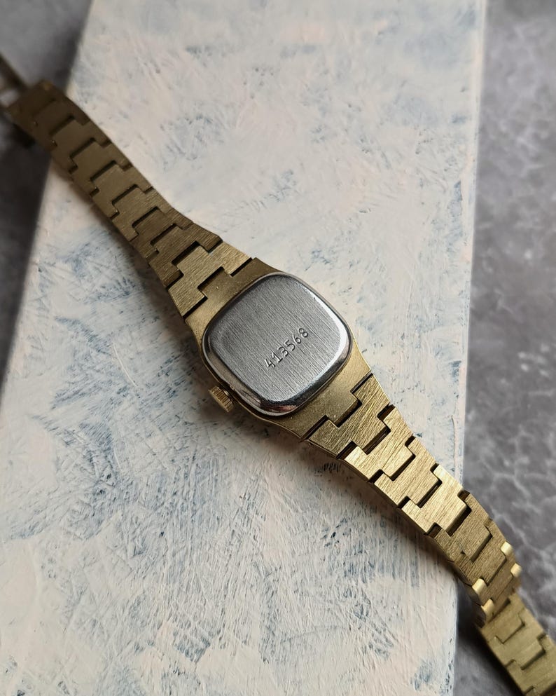 Pu&ograve; includere: Un orologio da polso vintage color oro con un quadrante rettangolare color argento. Il cinturino dell'orologio &egrave; composto da maglie in metallo color oro. L'orologio ha il numero "413568" inciso sul retro. L'orologio &egrave; posto su uno sfondo chiaro e strutturato.