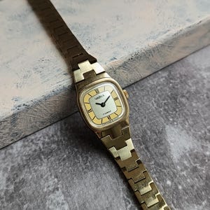 Pu&ograve; includere: Un orologio da polso vintage color oro con quadrante rettangolare e cinturino a maglie abbinato. Il quadrante &egrave; giallo chiaro con numeri romani neri e la parola cirillica "ЧАЙКА". L'orologio &egrave; posto su uno sfondo strutturato.