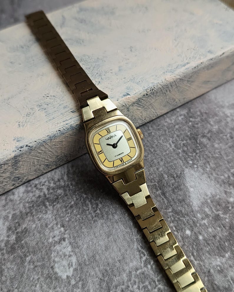 Pu&ograve; includere: Un orologio da polso vintage color oro con quadrante rettangolare e cinturino a maglie abbinato. Il quadrante &egrave; giallo chiaro con numeri romani neri e la parola cirillica "ЧАЙКА". L'orologio &egrave; posto su uno sfondo strutturato.