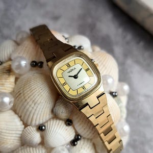 Pu&ograve; includere: Un orologio da polso vintage color oro con quadrante quadrato e cinturino in metallo abbinato. Il quadrante dell'orologio &egrave; giallo chiaro con lancette nere e numeri romani. L'orologio &egrave; etichettato "ЧАЙКА" e "17 KAMHEЙ".