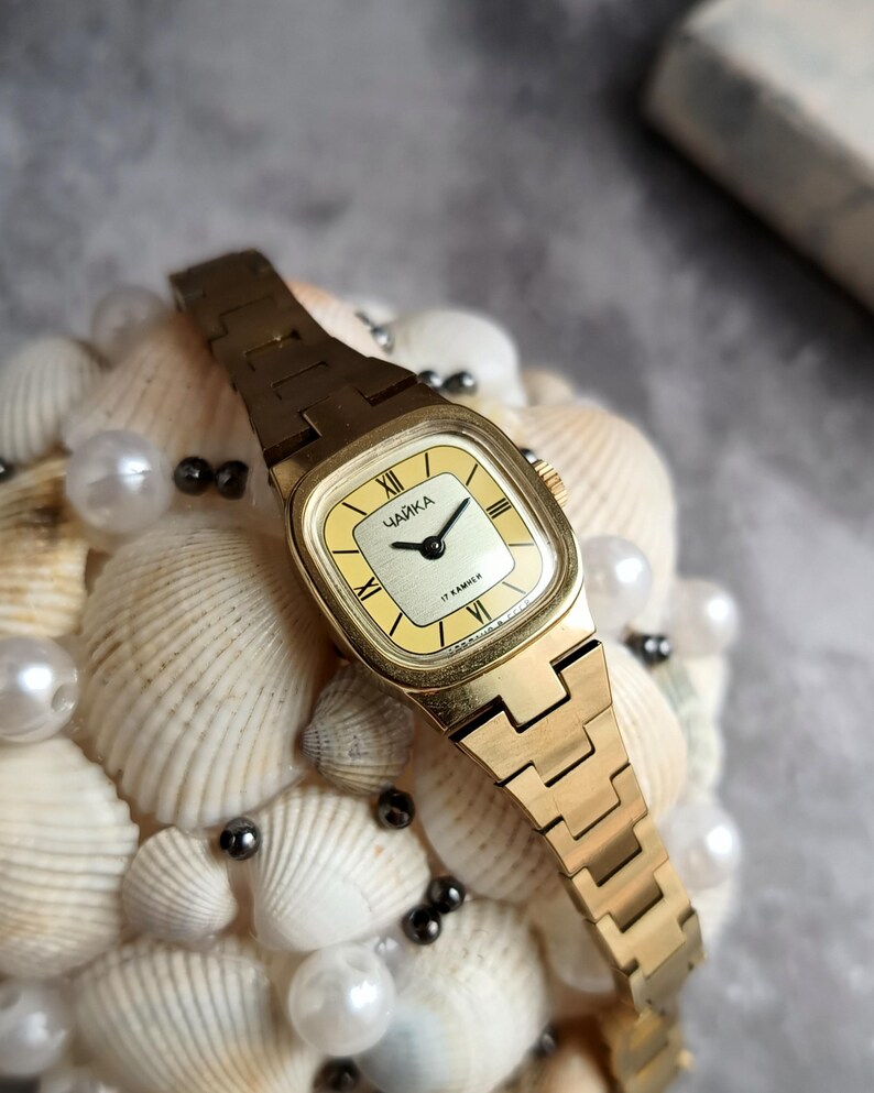 Pu&ograve; includere: Un orologio da polso vintage color oro con quadrante quadrato e cinturino in metallo abbinato. Il quadrante dell'orologio &egrave; giallo chiaro con lancette nere e numeri romani. L'orologio &egrave; etichettato "ЧАЙКА" e "17 KAMHEЙ".