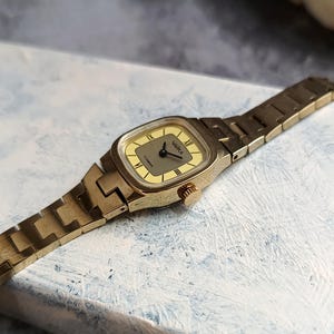 Pu&ograve; includere: Un orologio da polso vintage color oro con quadrante rettangolare e cinturino a maglie abbinato. Il quadrante dell'orologio &egrave; di colore giallo chiaro con lancette nere e numeri romani. La parola "ЧАЙКА" &egrave; stampata sul quadrante.
