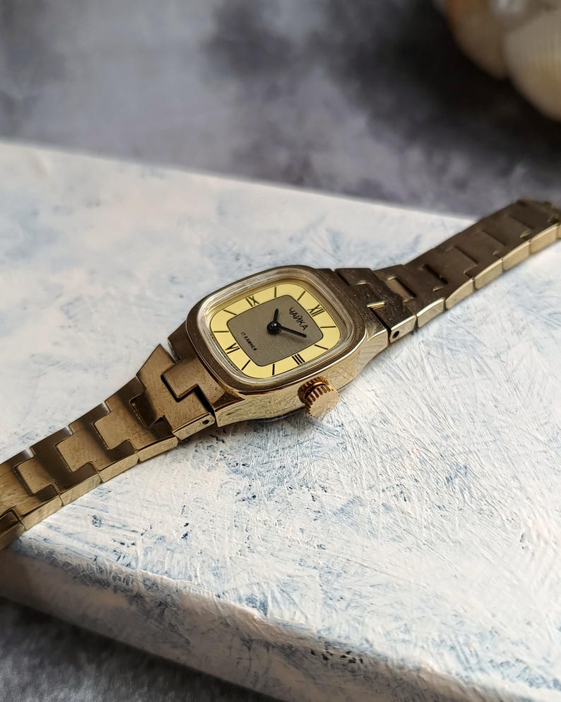 Pu&ograve; includere: Un orologio da polso vintage color oro con quadrante rettangolare e cinturino a maglie abbinato. Il quadrante dell'orologio &egrave; di colore giallo chiaro con lancette nere e numeri romani. La parola "ЧАЙКА" &egrave; stampata sul quadrante.