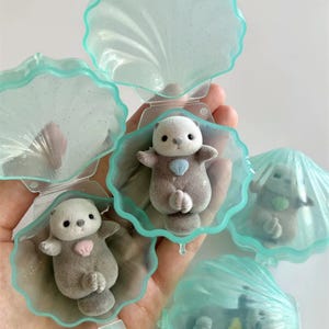 May include: A collection of miniature otter figurines, each nestled inside a translucent, light blue seashell-shaped container. The otters are primarily gray with white faces and small heart-shaped accents. The shells have a glittery finish.