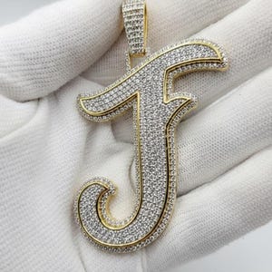 Puede incluir: Un colgante dorado con la forma de la letra "J", incrustado con numerosas piedras preciosas pequeñas y transparentes. El colgante está sobre un fondo blanco. Los bordes de la letra están delineados en oro. El colgante mide aproximadamente 5 cm de alto.