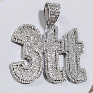 May include: A silver pendant spelling out "3tt" encrusted with small, sparkling stones. The pendant has a bail for a chain and is set against a white background. The letters are in a bold, stylized font.