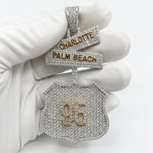 May include: A diamond-encrusted pendant in the shape of a shield with the number "96" in gold. Two intersecting signs read "CHARLOTTE" and "PALM BEACH" in gold lettering. The pendant is held against a white background.